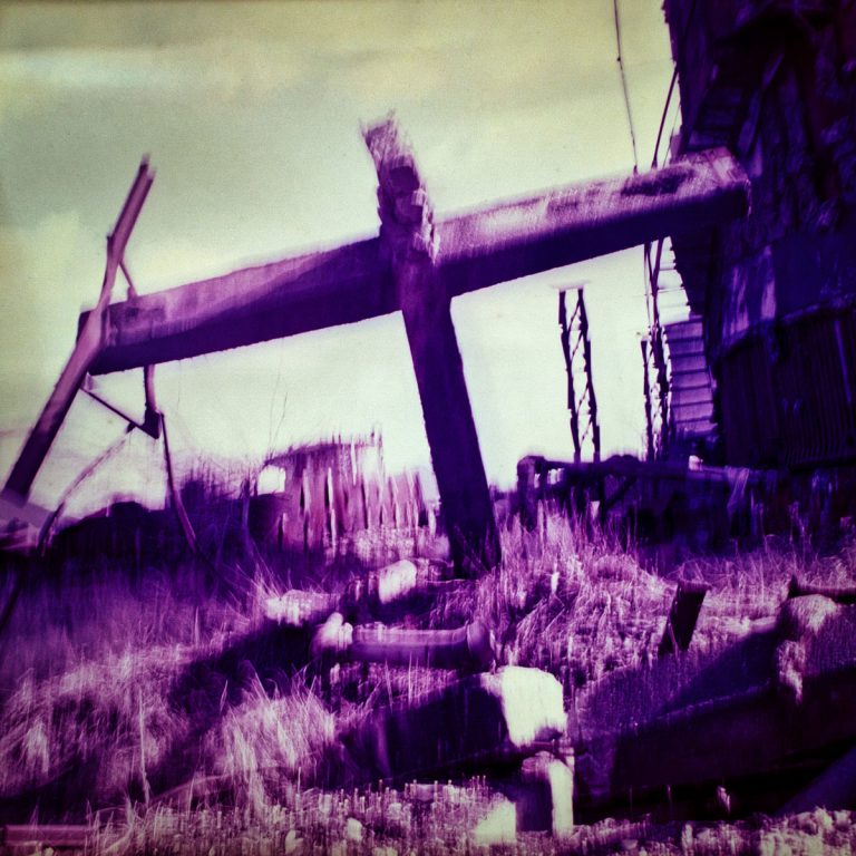 Contemporary fine art photograph shot on slide film by István Havadi-Nagy-David. Showing a collapsed industrial beam, resembling a crucifix, amidst overgrown grasses in a decaying landscape. The weathered structure leans at an unnatural angle, symbolizing the decline of human industry. A rusted factory or silo with a spiral staircase looms in the background. The surreal color palette of purples and yellows, along with blurred details, creates a dreamlike, distorted atmosphere. The artwork explores themes of abandonment, decay, and the quiet reclamation of industrial spaces by nature. The fourth image from the Postindustrial Golgotha series.
