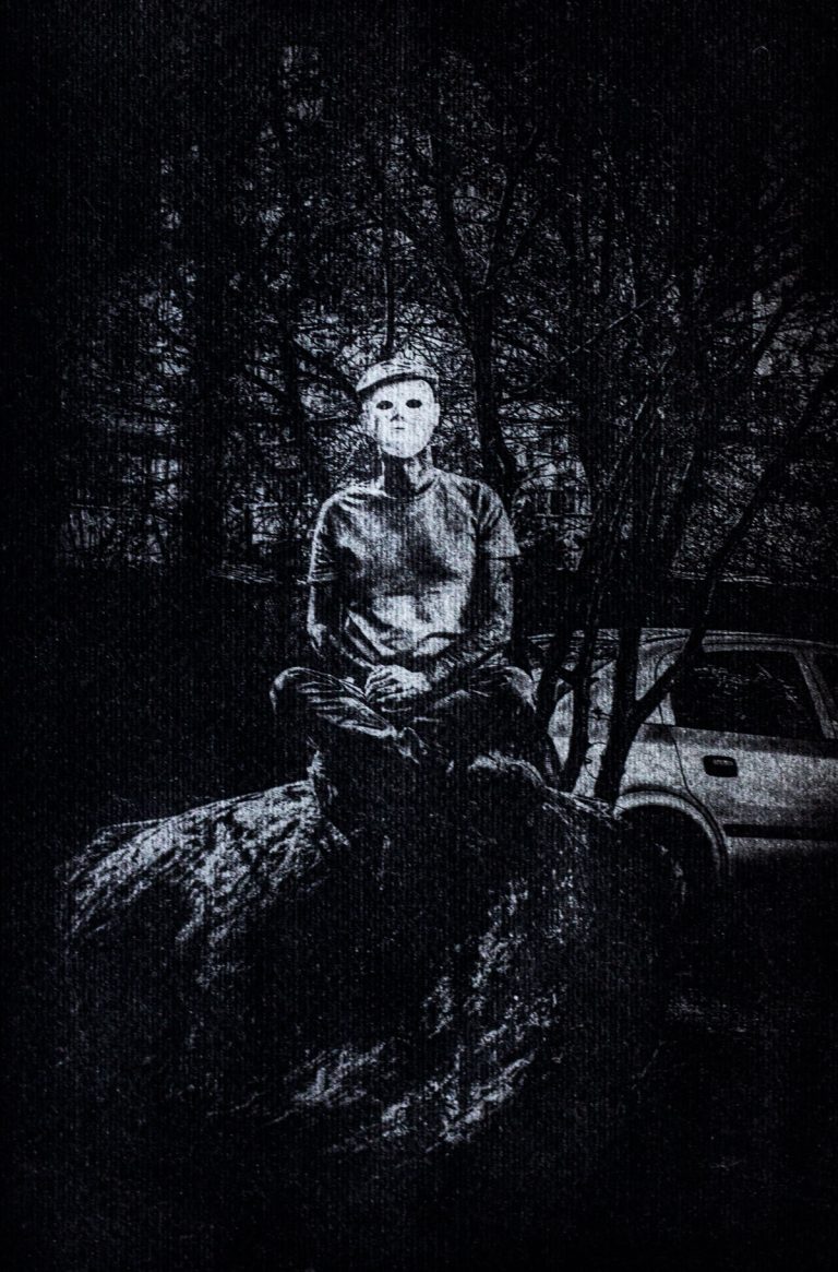 Unique fine art photograph by István Havadi-Nagy-David, printed with the gumoil technique. It is a dark, monochrome image that depicts a figure wearing a blank, white mask, sitting cross-legged on a large, uneven rock. The figure, dressed in casual clothes and a hat with an expressionless mask, appears to be in a meditative pose. The background is shadowy, with barren trees and the faint outline of a car visible to the right. The scene feels eerie and unsettling, with stark contrasts between light and shadow.